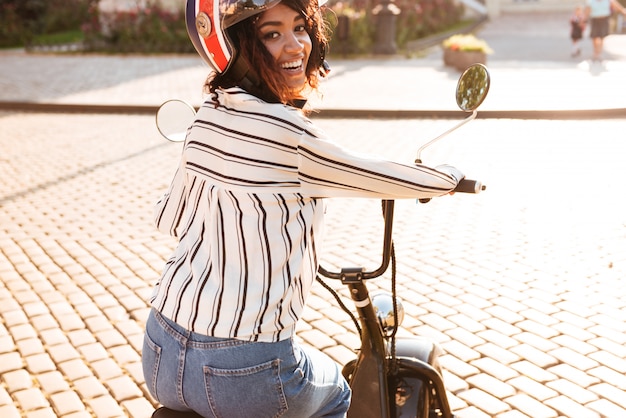 Kostenloses Foto hintere ansicht der freudigen afrikanischen frau in moto halmet reitet auf modernem motorrad im freien und betrachtet die kamera