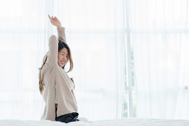 Hintere Ansicht der glücklichen schönen jungen asiatischen Frau, die am Morgen aufwacht und auf Bett sitzen