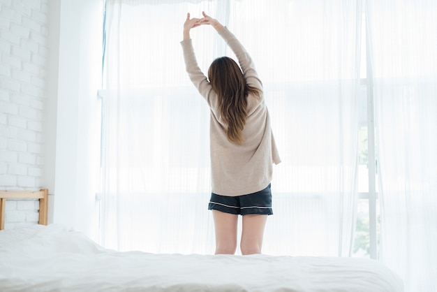 Hintere Ansicht der glücklichen schönen jungen asiatischen Frau, die am Morgen aufwacht und auf Bett sitzen