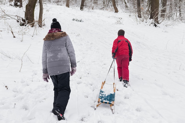 Hintere Ansicht des Jungen und des Mädchens, die mit leerem Schlitten in schneebedeckten Wald gehen