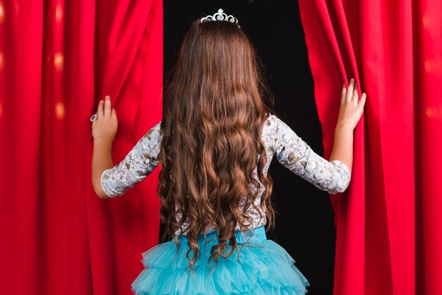 Hintere Ansicht des Mädchens mit dem gewellten Haar des langen Brunette, das vom roten Vorhang schaut