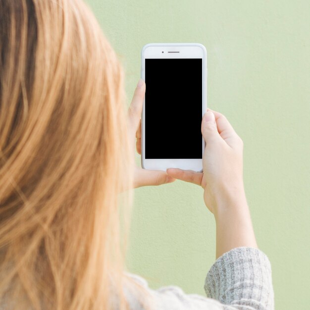 Hintere Ansicht einer blonden jungen Frau, die Handy gegen tadellosen grünen Hintergrund hält