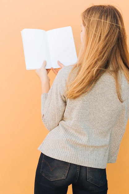 Hintere Ansicht einer blonden jungen Frau, die in der Hand Weißbuch gegen Pfirsichhintergrund hält