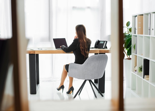 Hintere Ansicht einer Geschäftsfrau, die an dem Laptop gesehen durch Glasfenster arbeitet