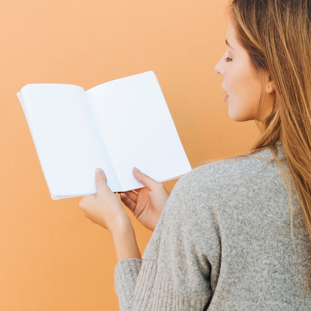 Kostenloses Foto hintere ansicht einer jungen frau, die in der hand weißbuch gegen pfirsichhintergrund hält