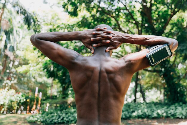 Hintere Ansicht eines hemdlosen jungen Mannes mit seinen Händen hinter dem Kopf, der in den Park ausdehnt