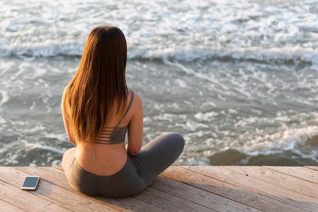 Hintere Ansicht junge Frau, die neben Meer meditiert