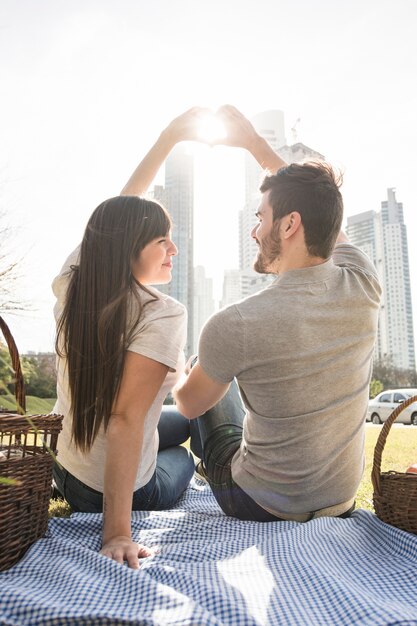 Hintere Ansicht von den jungen Paaren, die Herz mit der Hand im Sonnenlicht am Picknick bilden