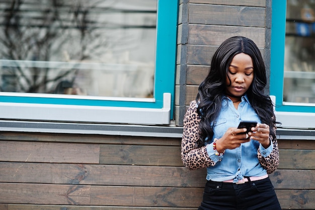 Hipster afroamerikanisches Mädchen mit Jeanshemd mit Leopardenärmeln hält Handy zur Hand und posiert auf der Straße gegen Holzhaus mit Fenstern