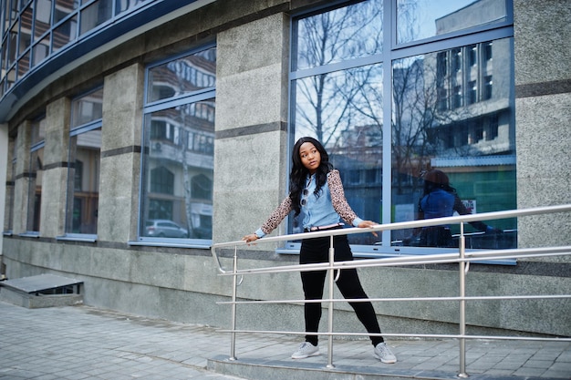 Hipster afroamerikanisches Mädchen mit Jeanshemd und Leopardenärmeln posiert auf der Straße vor einem modernen Bürogebäude mit blauen Fenstern