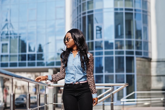 Hipster afroamerikanisches Mädchen mit Sonnenbrille Jeanshemd mit Leopardenärmeln posiert auf der Straße vor einem modernen Bürogebäude mit blauen Fenstern