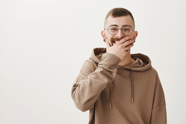 Hipster in Hoodie und Brille lachen, Mund mit der Hand bedecken, während kichern