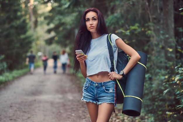 Hipster-Mädchen in Hemd und Shorts mit Touristenmatte und Rucksack hält ein Smartphone und schaut weg, während es in einem Sommerwald steht.