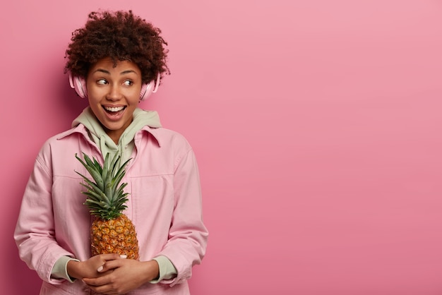 Hipster-mädchen mit freudigem ausdruck, breitem lächeln, in hochstimmung, genießt freizeit, hört lieblingssong über stereokopfhörer, posiert mit exotischer ananas, trägt hoodie, schaut zur seite