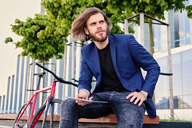 Hipster-Männchen mit langen blonden Haaren spricht per Smartphone und hält Tablet-PC mit Single-Speed-Fahrrad im Hintergrund.