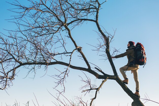 Hipster Mann, der mit Rucksack reist, auf Baum gegen Himmel steht, warme Jacke trägt, aktiver Tourist, Natur in der kalten Jahreszeit erforschend
