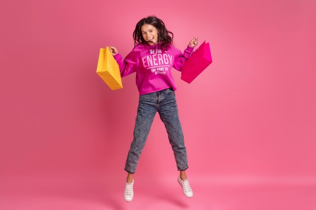 Hispanische hübsche Frau in rosa Hoodie und Jeans lächelnd auf rosa isoliert springend mit bunten Einkaufstüten Verkaufsrabatt