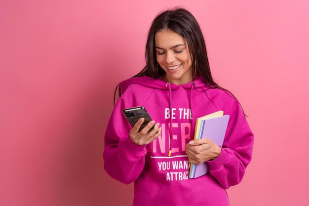 Hispanische hübsche Frau in rosafarbenem Hoodie lächelt, hält Notizbücher in der Hand und benutzt Smartphone