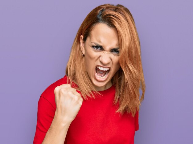 Hispanische junge Frau, die lässiges rotes T-Shirt trägt, wütend und wütend die Faust erhebt, frustriert und wütend, während sie vor Wut schreit. Wut und aggressives Konzept.