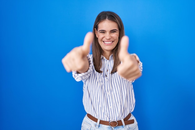 Hispanische junge Frau steht vor blauem Hintergrund und macht zustimmend eine positive Geste mit der Hand, Daumen hoch, lächelnd und glücklich über den Erfolg. Siegergeste.