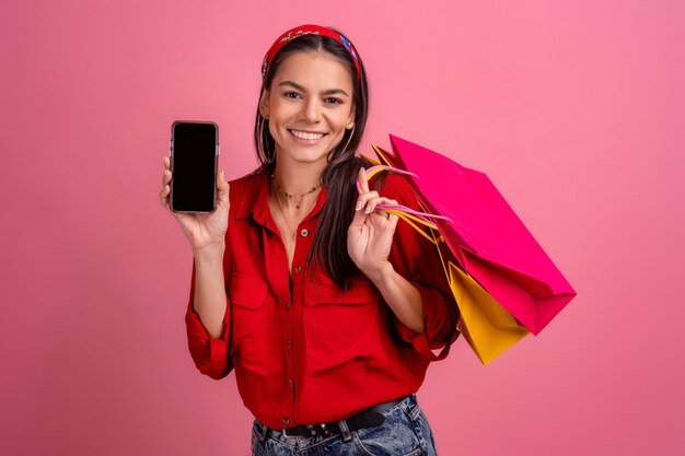Hispanische schöne Frau im roten Hemd, die lächelnd hält und Einkaufstaschen und Smartphone auf rosa Studiohintergrund hält, isolierter Rabattverkauf schwarzer Freitag