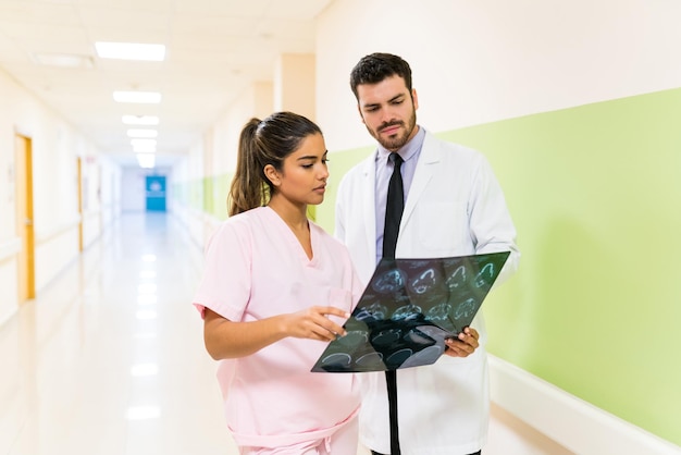 Hispanischer Arzt und Krankenschwester diskutieren über Röntgenaufnahmen, während sie zusammen im Korridor des Krankenhauses stehen