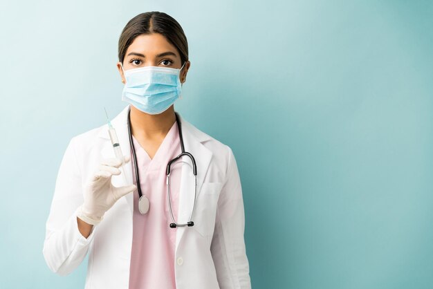 Hispanischer junger Arzt, der eine Spritze hält, während er eine Maske vor blauem Hintergrund trägt