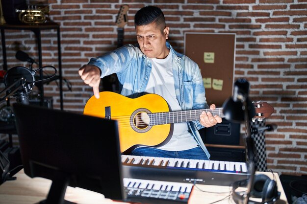 Hispanischer junger Mann, der im Musikstudio klassische Gitarre spielt, sieht unglücklich und wütend aus und zeigt Ablehnung und negative Geste mit schlechtem Gesichtsausdruck