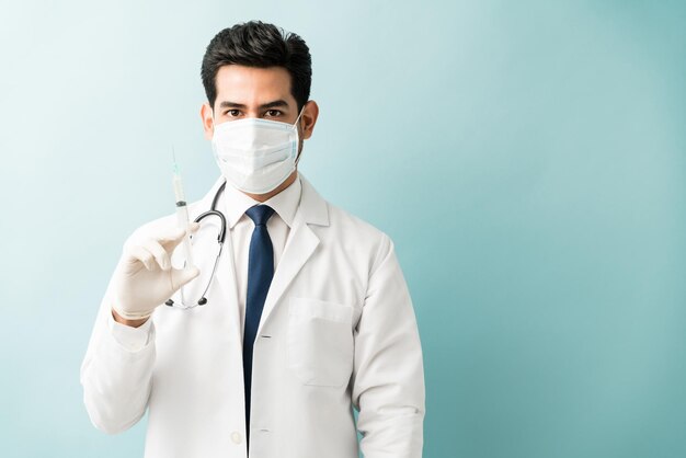 Hispanischer männlicher Arzt mit Spritze, während er Maske vor isoliertem Hintergrund trägt