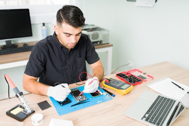 Hispanischer männlicher Ingenieur, der in seiner Reparaturwerkstatt die Anschlüsse eines beschädigten Smartphones mit einem Multimeter überprüft