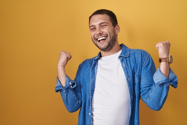 Hispanischer Mann, der vor gelbem Hintergrund steht und mit erhobenen Armen und geschlossenen Augen überrascht und erstaunt über den Erfolg feiert. Gewinner-Konzept.