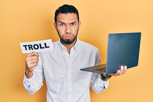 Hispanischer Mann mit Bart, der mit einem Computer-Laptop arbeitet und ein Banner mit Trollwort hält, das Wangen mit lustigem Gesicht aufbläst. Mund mit Luft aufgeblasen, Luft fangend.