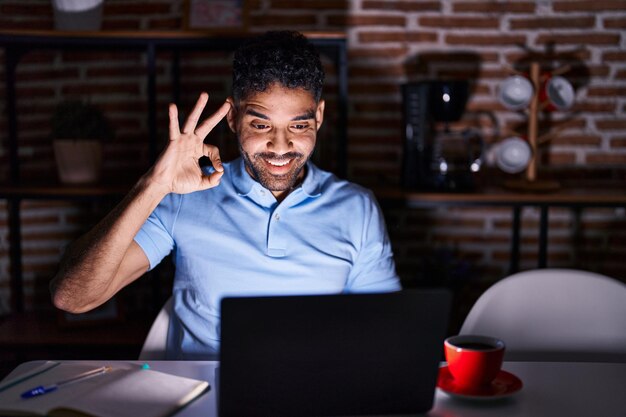 Hispanischer Mann mit Bart, der nachts einen Laptop benutzt, lächelt positiv und macht mit Hand und Fingern ein gutes Zeichen mit erfolgreichem Gesichtsausdruck