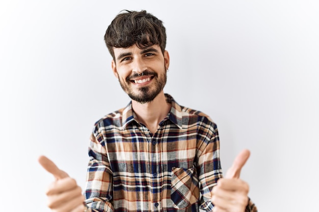 Hispanischer Mann mit Bart, der vor isoliertem Hintergrund steht und positive Gesten mit erhobenem Handdaumen lächelnd und glücklich über den Erfolg als Siegergeste genehmigt