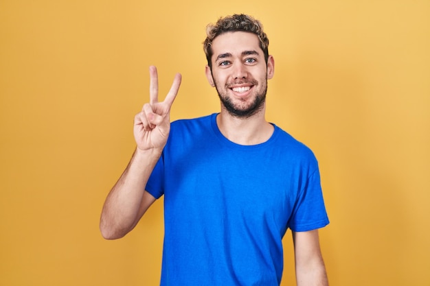 Hispanischer Mann mit Bart steht vor gelbem Hintergrund und lächelt mit glücklichem Gesicht, zwinkert in die Kamera und macht mit den Fingern ein Siegeszeichen. Nummer zwei.