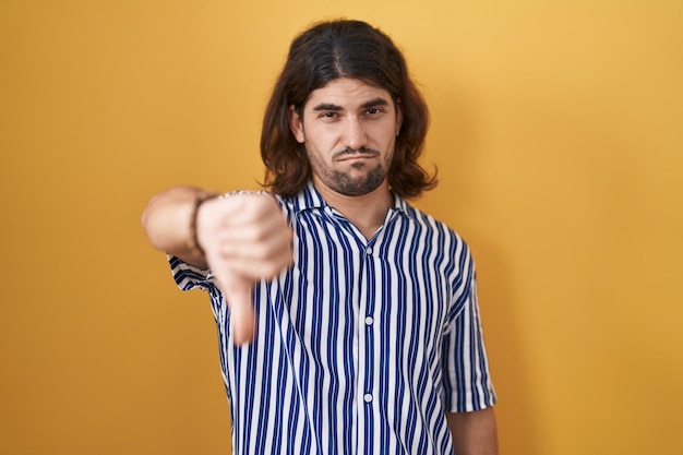 Kostenloses Foto hispanischer mann mit langen haaren, der über gelbem hintergrund steht und unglücklich und wütend aussieht und ablehnung und negativ mit daumen nach unten zeigt. schlechter ausdruck.