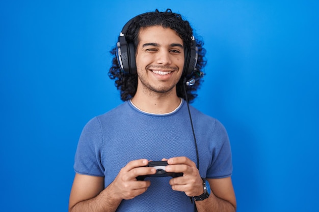 Hispanischer Mann mit lockigem Haar, der ein Videospiel spielt und einen Controller hält, der mit sexy Ausdruck, fröhlichem und glücklichem Gesicht in die Kamera schaut.
