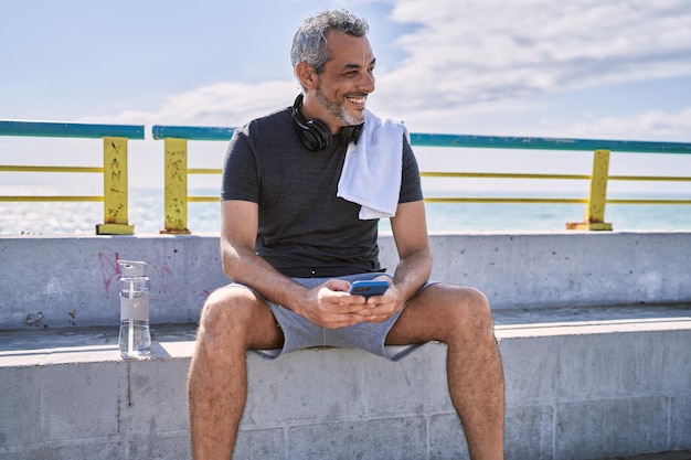 Hispanischer Mann mittleren Alters trägt Sportkleidung und nutzt sein Smartphone am Meer