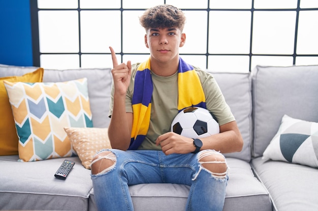 Hispanischer Teenager sitzt auf dem Sofa und schaut sich ein Fußballspiel an, lächelt glücklich und zeigt mit der Hand und dem Finger zur Seite