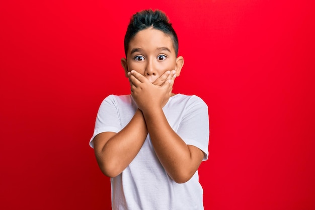 Hispanisches Kind des kleinen Jungen, das lässiges weißes T-Shirt trägt, schockiert, Mund mit den Händen für Fehler bedeckend. geheimes Konzept.