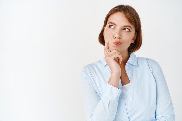 Hmm, interessant. Porträt eines nachdenklichen jungen Mädchens, das beiseite schaut und nachdenkt, denkt oder einen Plan macht, in Bluse auf weißer Wand steht standing