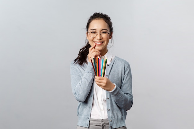 Hobbys, Kreativität und Kunstkonzept. Dumme glücklich lächelnde asiatische Frau, Künstlerin in Brille kichert als mit Buntstiften, hat interessante Idee für ihr nächstes Kunstwerk, Zeichnungsbild, grauer Hintergrund.