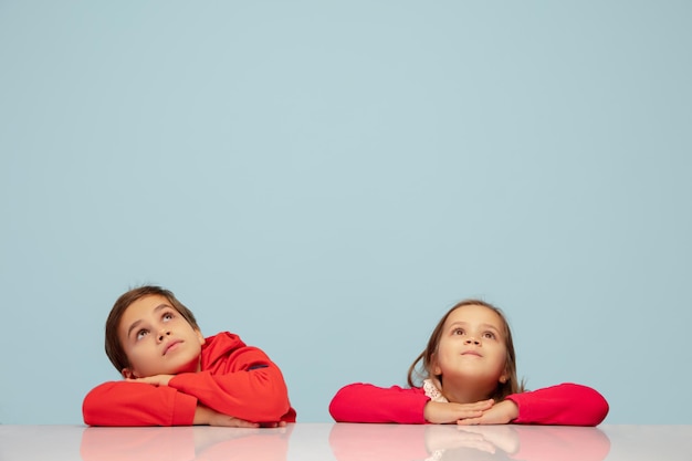 Hoch schauen. Glückliche Kinder lokalisiert auf blauem Studiohintergrund. Schauen Sie glücklich, fröhlich. Exemplar für Anzeige. Kindheit, Bildung, Emotionen, Gesichtsausdruckkonzept. Traumhaft lächelnd am Tisch sitzen