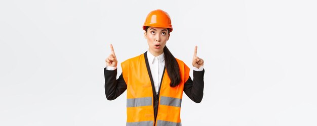 Hochbau und Industriekonzept Überrascht und neugierig asiatische Bauleiterin in Schutzhelm und reflektierender Kleidung, die mit dem Finger auf ein interessantes Banner zeigt