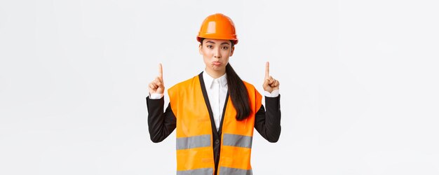 Hochbau und Industriekonzept Unzufriedene traurige asiatische Architektin, die sich beschwert, einen Schutzhelm und reflektierende Kleidung zu tragen, die verärgert ist, als sie mit dem Finger nach oben zeigt
