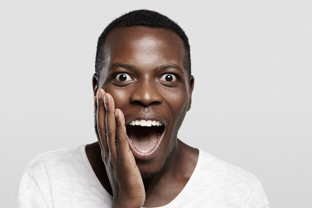 Hochdetailliertes Nahaufnahmeporträt eines aufgeregten afrikanischen Mannes im weißen T-Shirt, das überrascht aussieht, vor Schock mit weit geöffnetem Mund schreit, Hand auf Wange hält, erstaunt über eine unglaubliche Geschichte
