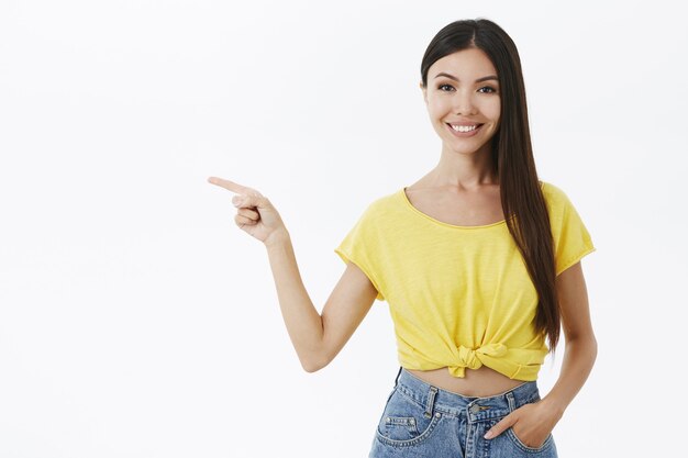 Höfliches und freundliches charmantes europäisches weibliches Modell mit langen dunklen Haaren im trendigen gelben kurz geschnittenen T-Shirt, das links zeigt und lächelt