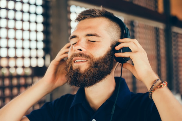 Hörende Musik des jungen Mannes in den Kopfhörern, unter Verwendung des Smartphone, Hippie-Porträt im Freien