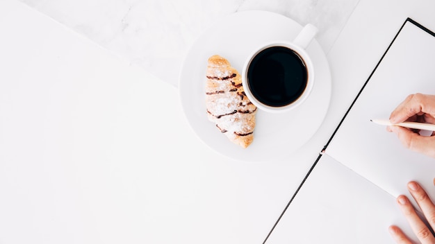 Hörnchen und Kaffeetasse auf Schreibtisch mit einem Personenschreiben im Tagebuch mit Bleistift