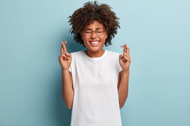 Hoffnungsvolle dunkelhäutige Frau mit zahnigem strahlendem Lächeln, trägt weißes T-Shirt, glaubt an Glück, hat Afro-Frisur, hofft, dass Träume wahr werden, isoliert über blauer Wand. Bitte, Gott hilf mir!